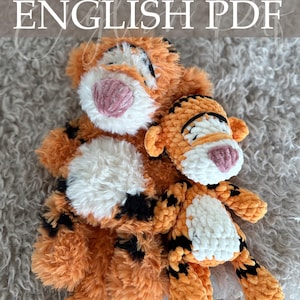 Op de afbeelding: Twee oranje en zwarte gehaakte tijgerknuffels met witte en roze accenten. De grotere tijger heeft een pluizige textuur, terwijl de kleinere een strakkere steek heeft. De tekst "Crochet Pattern ENGLISH PDF" is zichtbaar.