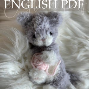 Op de afbeelding: Een zachte grijze en witte kattenknuffel met een roze bolletje wol. De kat zit op een zachte, witte ondergrond. De afbeelding bevat de tekst "Crochet Pattern ENGLISH PDF" en de naam van de ontwerper.