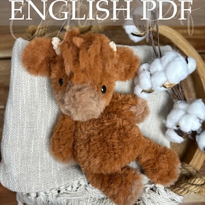 May include: A plush, brown Highland cow stuffed animal with small horns, resting on a light-colored fabric. The image includes the text "Crochet Pattern ENGLISH PDF" and is set against a rustic wooden background with cotton accents.
