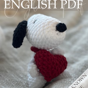 Op de afbeelding: Een gehaakte Snoopy-achtige hond met een rood hart. De hond is wit met zwarte oren en neus. De afbeelding bevat de tekst "Crochet Pattern ENGLISH PDF" en "WELL KNOWN SERIE".