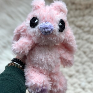 May include: A plush toy made of soft, fuzzy light pink yarn with long ears. It features two large, round black eyes with small white highlights and a textured, light purple nose. The creature has a rounded head and a soft, shaggy body texture.