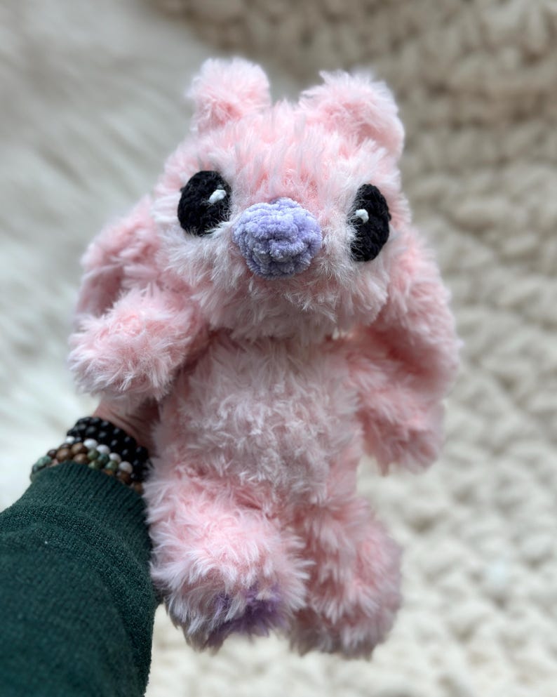 May include: A plush toy made of soft, fuzzy light pink yarn with long ears. It features two large, round black eyes with small white highlights and a textured, light purple nose. The creature has a rounded head and a soft, shaggy body texture.