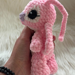 May include: A small, handmade crochet plush toy made of soft, light pink yarn. The creature features long, drooping ears, a small purple nose, and large, round black eyes with white accents. The texture is fuzzy and plush, with a rounded head and body.