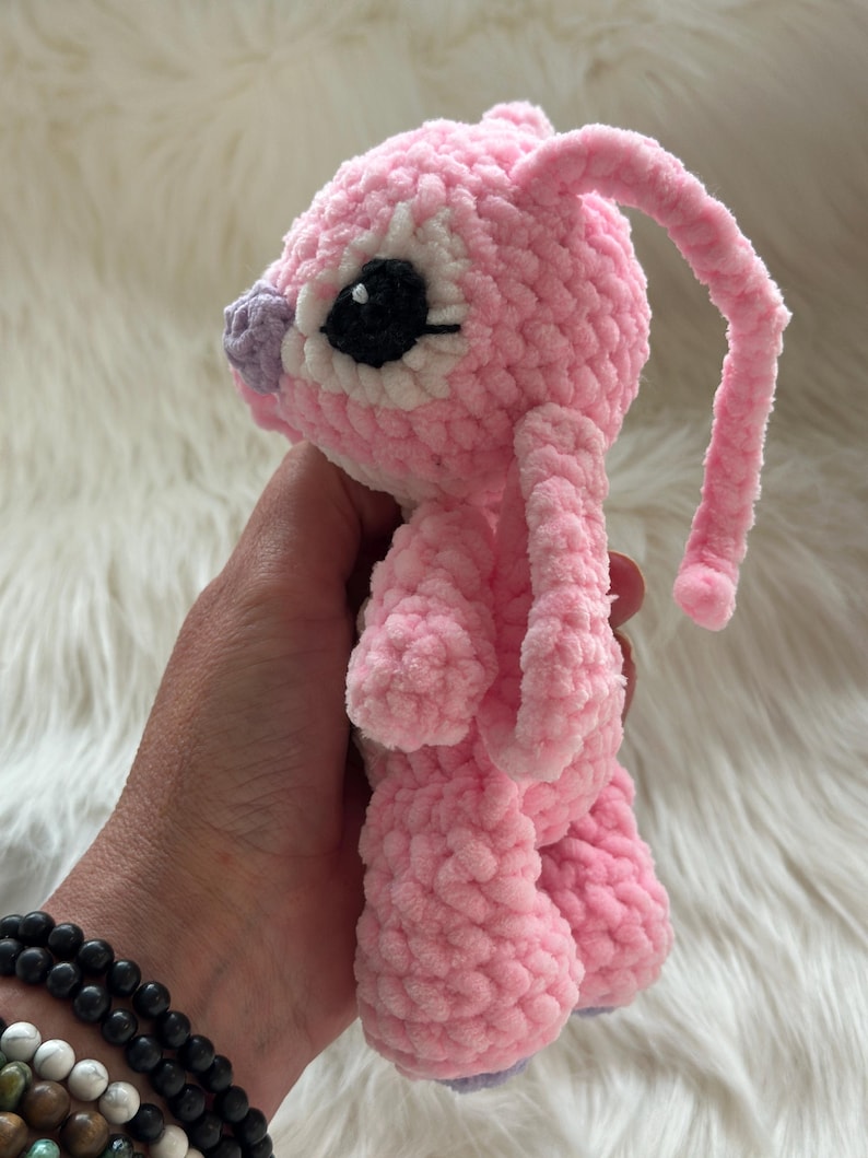 May include: A small, handmade crochet plush toy made of soft, light pink yarn. The creature features long, drooping ears, a small purple nose, and large, round black eyes with white accents. The texture is fuzzy and plush, with a rounded head and body.