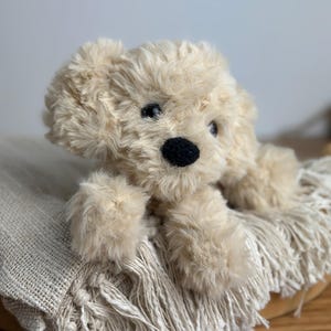 May include: A plush, cream-colored dog toy with a fluffy texture. The toy has a black nose and dark eyes, and is lying on a fringed, cream-colored blanket. The dog's fur is soft and shaggy, giving it a cuddly appearance.