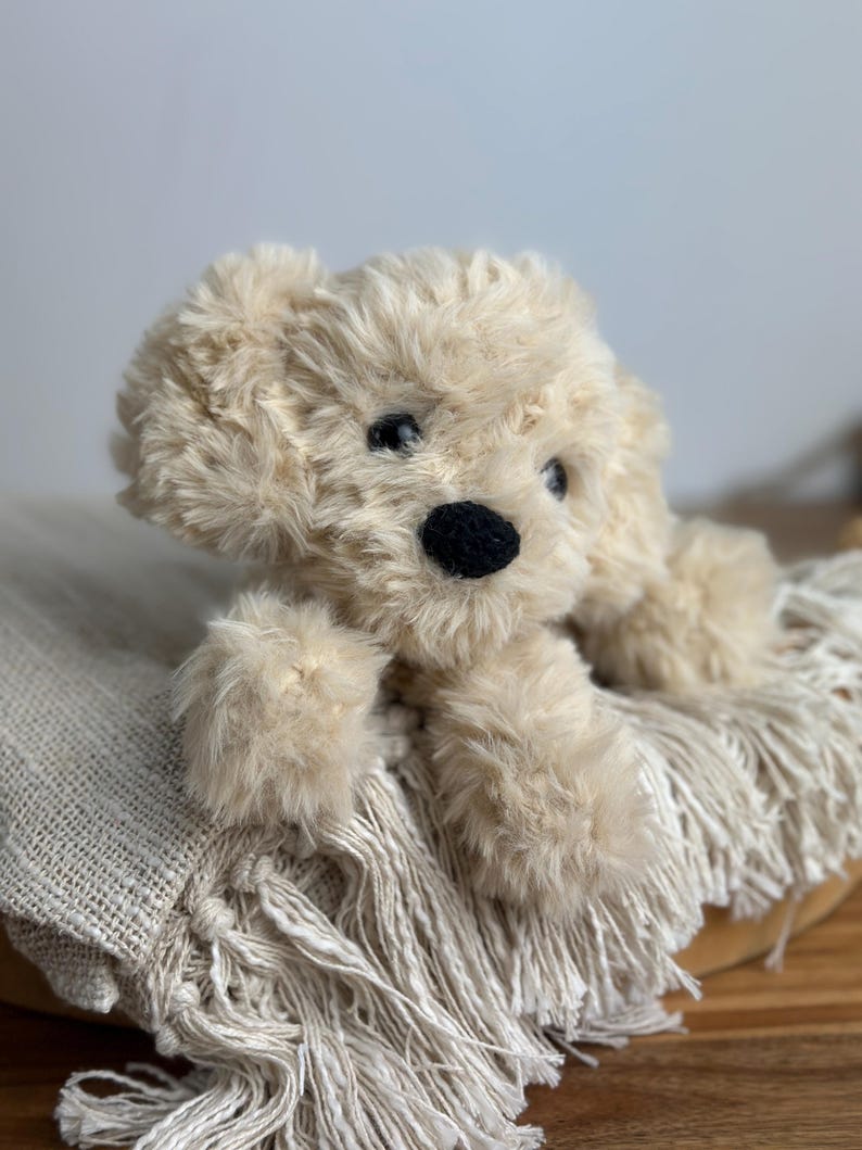 May include: A plush, cream-colored dog toy with a fluffy texture. The toy has a black nose and dark eyes, and is lying on a fringed, cream-colored blanket. The dog's fur is soft and shaggy, giving it a cuddly appearance.