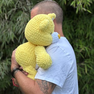 K&ouml;nnte beinhalten: Ein handgefertigter, gelber H&auml;kelteddyb&auml;r mit einem wei&szlig;en und gelben Blumenakzent. Der B&auml;r wird gehalten. Die weiche Textur und die leuchtende Farbe des B&auml;ren sind sichtbar. Der B&auml;r ist ein kuscheliges Spielzeug.