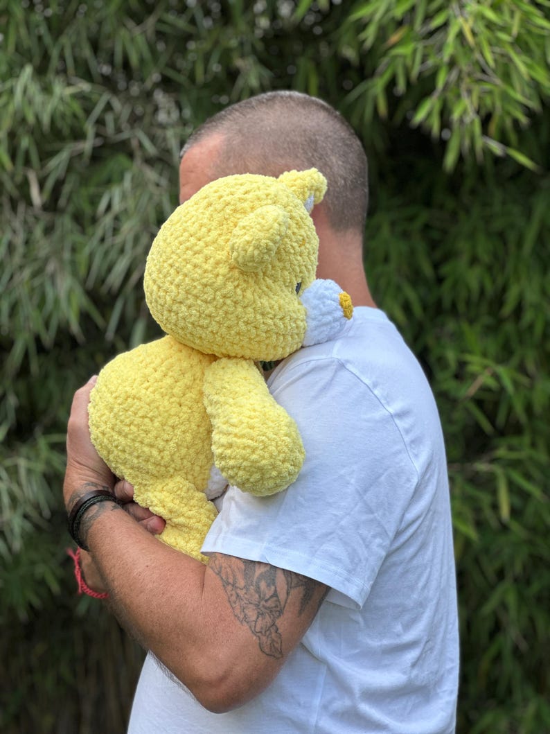 K&ouml;nnte beinhalten: Ein handgefertigter, gelber H&auml;kelteddyb&auml;r mit einem wei&szlig;en und gelben Blumenakzent. Der B&auml;r wird gehalten. Die weiche Textur und die leuchtende Farbe des B&auml;ren sind sichtbar. Der B&auml;r ist ein kuscheliges Spielzeug.