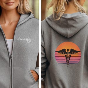 May include: A gray zip-up hoodie with the name "Samantha, RN" embroidered on the front. The back features a sunset design with a medical caduceus symbol in black. The hoodie has a hood and front pockets.