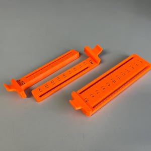 Puede incluir: Dos reglas de plástico naranja con marcas negras. La regla de la izquierda tiene marcas del 11 al 15J y la regla de la derecha tiene marcas del 15 al 18 y FLE.