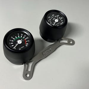 Könnte beinhalten: Zwei schwarze Motorradanzeigen mit weißen Markierungen und Zeigern sowie eine silberne Metallhalterung. Ein Messgerät zeigt die Geschwindigkeit bis zu 140 mph an, das andere die Drehzahl. Auf der Halterung sind die Buchstaben "ZNC" eingraviert.