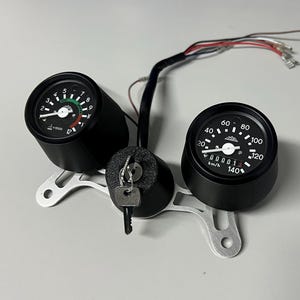 May include: Black motorcycle gauge cluster with two round gauges, a key ignition, and wiring. The gauges are mounted on a silver bracket. One gauge displays RPM, the other displays speed in km/h.