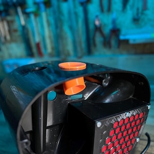 May include: A black plastic device with an orange nozzle and a red honeycomb filter. The device has a curved top and appears to be a component of a larger machine or tool. The background is a blurred workshop setting.