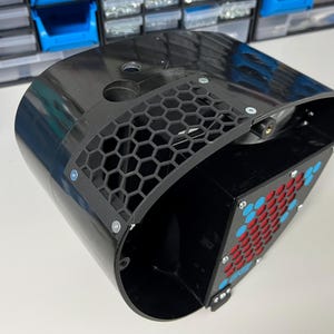 May include: A black plastic fan housing with a honeycomb-shaped grill on the side. The fan is visible inside the housing, and there are red and blue accents on the grill.
