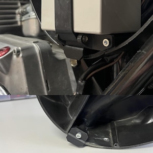 May include: Close-up of motorcycle components, including a black bracket and a silver engine part. The image shows a black strap securing a rectangular object. The components are set against a dark background, highlighting the details of the motorcycle.