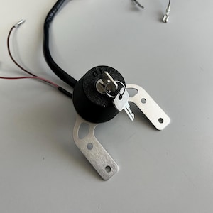 May include: A black ignition switch with two keys inserted. The switch is mounted on a silver metal bracket with two holes. Wires extend from the switch. The item is likely an automotive or motorcycle part.