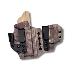 Op de afbeelding: Een camouflage-patroon holster met zwarte clips en hardware. De holster heeft een getextureerd oppervlak en een lichtbruine achterkant. Het ontwerp omvat meerdere bevestigingspunten en een veilig clipsysteem voor het dragen van een vuurwapen.