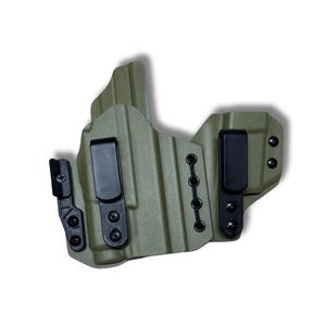 Op de afbeelding: Een olijfgroene, gegoten polymeer holster met twee zwarte riemclips en een reeks oogjes voor verstelbare retentie. De holster is ontworpen om in de tailleband te worden gedragen en heeft een getextureerd oppervlak.