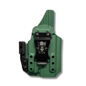 Op de afbeelding: Een olijfgroene gevormde holster met een zwarte metalen riemclip. De clip toont een witte afbeelding van een schedel met een hoge hoed en de tekst Voodoo Armory. Een zwart getextureerd hulpstuk is aan de zijkant bevestigd met drie schroeven.