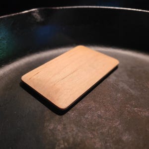 May include: A small, rectangular wooden cutting board with rounded corners rests inside a black cast iron skillet. The cutting board is light brown with a visible wood grain and a crack running through the center.