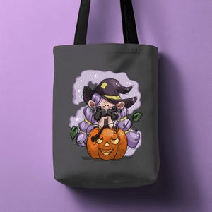 Peut inclure: Un sac fourre-tout gris foncé avec une illustration sur le thème d'Halloween. Le motif comprend une sorcière de dessin animé aux cheveux violets et un chapeau de sorcière, tenant une citrouille orange sculptée. Des feuilles vertes et un fond violet en forme de nuage complètent le design.
