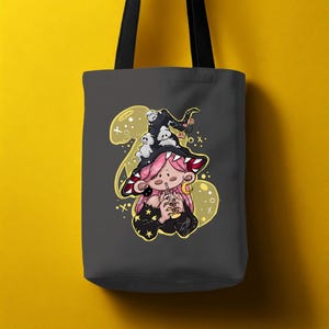 May include: A dark grey tote bag with a cartoon illustration of a witch character with pink hair, wearing a black hat with white ghosts. The witch is holding a drink with a straw. The background has yellow accents.