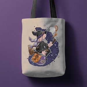 May include: Beige tote bag with a cartoon witch illustration. The witch, wearing a black hat and coat, rides a broomstick against a starry backdrop. The bag features black straps and a keyhole design on the bag.
