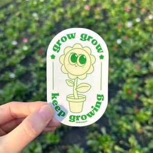 Peut inclure: Un autocollant blanc avec un contour et du texte verts. L'autocollant représente une fleur de dessin animé avec un visage souriant dans un pot. Les mots "grow grow" et "keep growing" sont écrits autour de la fleur. L'autocollant est tenu par une main.