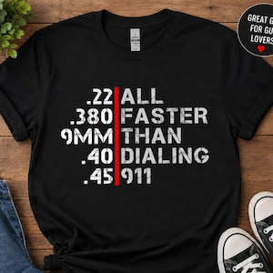 Peut inclure: Un t-shirt noir à manches courtes avec un texte blanc vieilli indiquant .22 .380 9MM .40 .45 ALL FASTER THAN DIALING 911, séparé par une ligne rouge verticale. Le t-shirt présente une encolure ronde et une coupe standard, posé à plat.