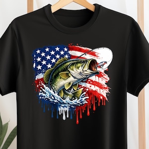May include: Black t-shirt featuring a graphic of a largemouth bass leaping from water, with a fishing lure and rod. The design incorporates the American flag with red, white, and blue colors, creating a patriotic theme. The shirt is hanging on a wooden hanger.