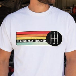 May include: A white t-shirt featuring a rectangular graphic with horizontal stripes in red, orange, yellow, and teal. The text "CLASSICALLY TRAINED" appears in black, next to a black circular gear shift pattern showing positions 1, 3, 5, 2, 4, and R.