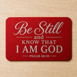 May include: A rectangular red metal plaque with rounded corners featuring silver engraved text that reads: Be Still and KNOW THAT I AM GOD PSALM 46:10. The lettering uses a combination of elegant script and bold serif fonts.