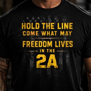 May include: Black t-shirt with a distressed American flag design and the text "HOLD THE LINE COME WHAT MAY FREEDOM LIVES IN THE 2A" in yellow. The shirt has a crew neck and short sleeves.