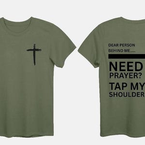 May include: Olive green t-shirt with a black cross on the front and back. The back of the shirt has the text "DEAR PERSON BEHIND ME... NEED PRAYER? TAP MY SHOULDER" in black block letters.