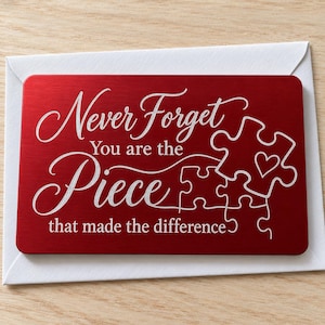 May include: A rectangular red metal card featuring white engraved text that reads Never Forget You are the Piece that made the difference, accompanied by a white puzzle piece graphic containing a small heart outline, resting on a white envelope.