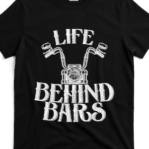 May include: A black short-sleeve t-shirt featuring a white graphic print of motorcycle handlebars and a headlight. The text "LIFE BEHIND BARS" is printed in a distressed white font above and below the motorcycle illustration on the chest.