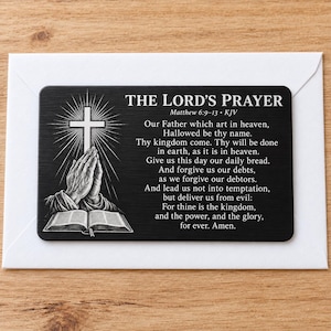 May include: A rectangular black metal card featuring the text THE LORD'S PRAYER Matthew 6:9-13 KJV. The design includes a white engraved illustration of praying hands, an open book, and a glowing cross above the prayer text in white lettering.