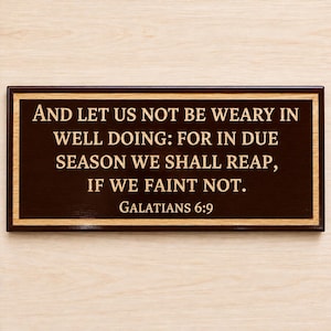 May include: A rectangular wooden wall plaque with a dark brown center and a light wood border. The text reads: AND LET US NOT BE WEARY IN WELL DOING: FOR IN DUE SEASON WE SHALL REAP, IF WE FAINT NOT. GALATIANS 6:9 in gold-colored lettering.