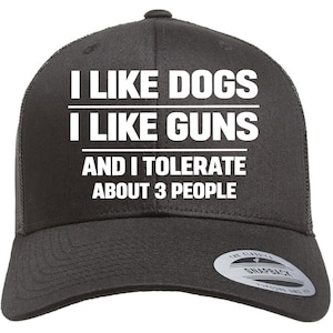 May include: Black trucker hat with white text that reads "I LIKE DOGS, I LIKE GUNS, AND I TOLERATE ABOUT 3 PEOPLE." The hat has a curved brim and a mesh back. A small tag is visible on the side.
