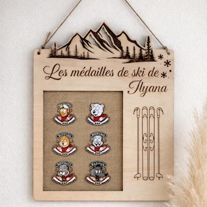 Peut inclure: Tableau en bois pour médailles de ski avec un motif de montagne et d'arbres. Le tableau a un fond en toile de jute et contient six épingles colorées sur le thème du ski. Le texte "Les médailles de ski de * Tlyana" est gravé sur le tableau.