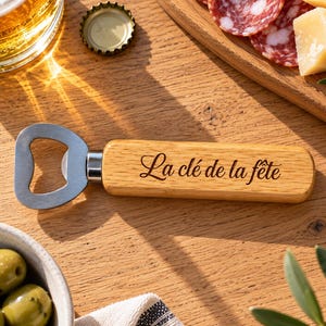 Puede incluir: Un abridor de botellas con mango de madera clara que presenta el texto grabado La clé de la fête. El cabezal metálico del abridor es de color plateado y está unido a la empuñadura rectangular de madera, con un acabado liso y bordes redondeados.