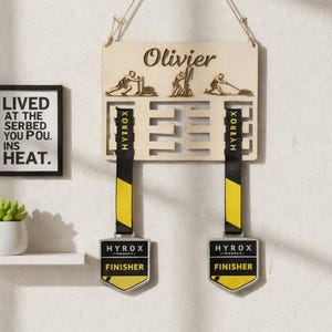 May include: Wooden medal display with the name "Olivier" and engraved athlete figures. It features slots for medals with black and yellow ribbons, each labeled "HYROX FINISHER." A framed print with text is on the left.