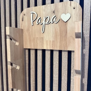 Könnte beinhalten: Ein helles Wandregal aus Holz mit einer flachen Rückwand, auf der das Wort Papa und ein Herz in weißer Schrift angebracht sind. Das Regal verfügt über zwei vertikale Seitenstützen mit jeweils drei eingekerbten Aussparungen zur Aufbewahrung.