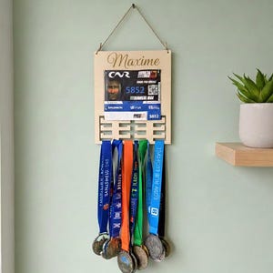 Peut inclure: Présentoir à médailles en bois avec le nom "Maxime" en écriture cursive. Le présentoir contient des dossards et plusieurs médailles avec des rubans colorés. Les médailles sont suspendues en dessous.