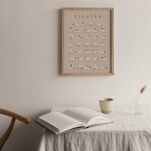 Peut inclure: Une affiche encadr&eacute;e avec le mot "PILATES" en haut et des illustrations de diff&eacute;rents exercices de pilates. L'affiche est accroch&eacute;e &agrave; un mur beige clair au-dessus d'une table avec un livre et une tasse.