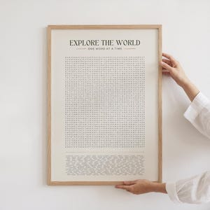 May include: A framed print with the text "EXPLORE THE WORLD" and "ONE WORD AT A TIME" in a minimalist style. The print features a word search puzzle in black against a cream background, encased in a light wood frame.
