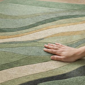 Pu&ograve; includere: Primo piano di un tappeto verde e beige con un motivo a onde. Il tappeto presenta varie tonalit&agrave; di verde, beige e un tocco di oro. Una mano &egrave; appoggiata sul tappeto, evidenziandone la consistenza. Il tappeto &egrave; probabilmente realizzato in un materiale morbido.