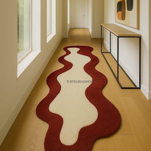 May include: A hallway runner rug with a unique, flowing design. The rug features a deep red border with a cream-colored center, creating a striking contrast. The rug is placed on a light wood floor, adding warmth to the space. The text "TEXTIELRUGSHOP" is visible on the rug.