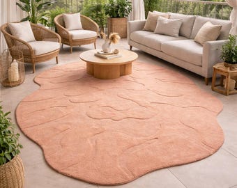 May include: A peach-colored, organically shaped rug dominates an outdoor living space. The rug is placed on a tiled floor, surrounded by wicker chairs, a sofa, and a wooden coffee table. The space is decorated with plants.