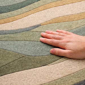 Pu&ograve; includere: Primo piano di un tappeto con un motivo a onde nei toni del verde, beige e grigio. Il tappeto ha un aspetto morbido e strutturato. Una mano &egrave; appoggiata sul tappeto, mostrando la consistenza e il tatto del materiale.